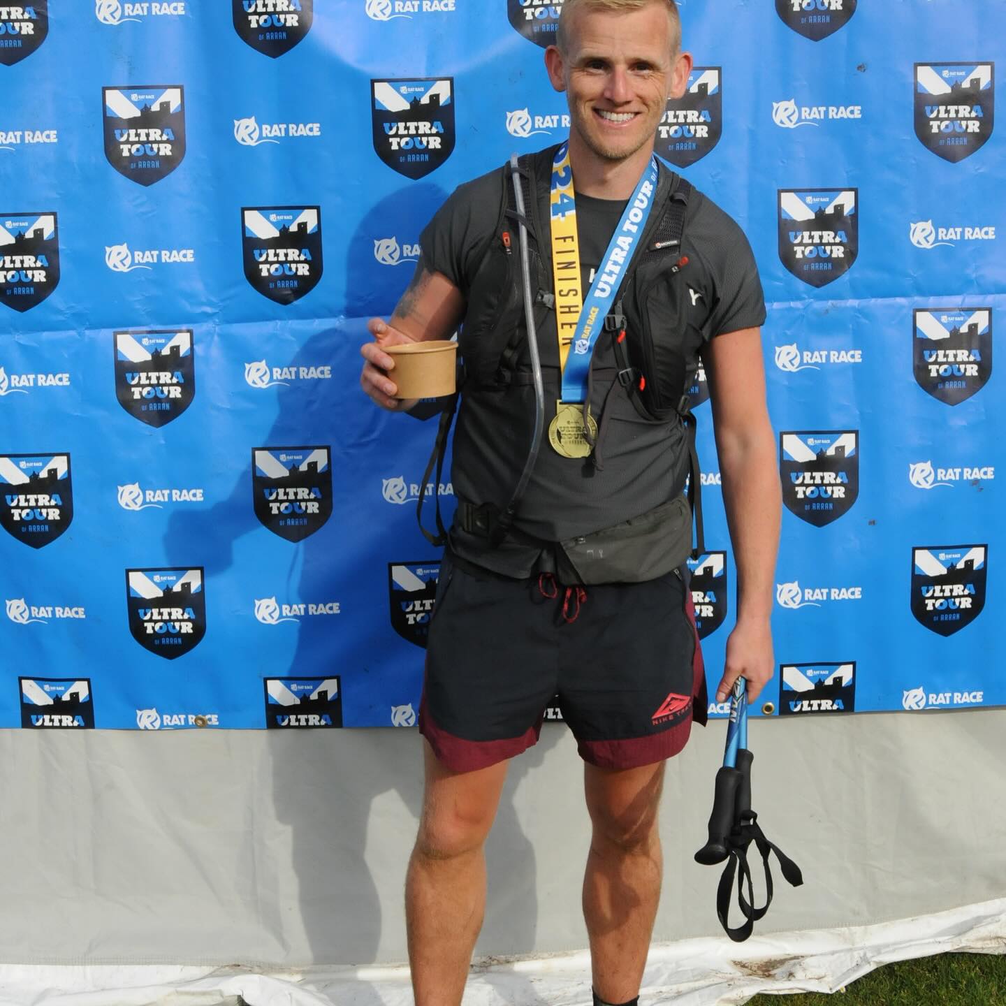 A runner wearing a finisher medal in front of the Rat Race Ultra Tour backdrop.