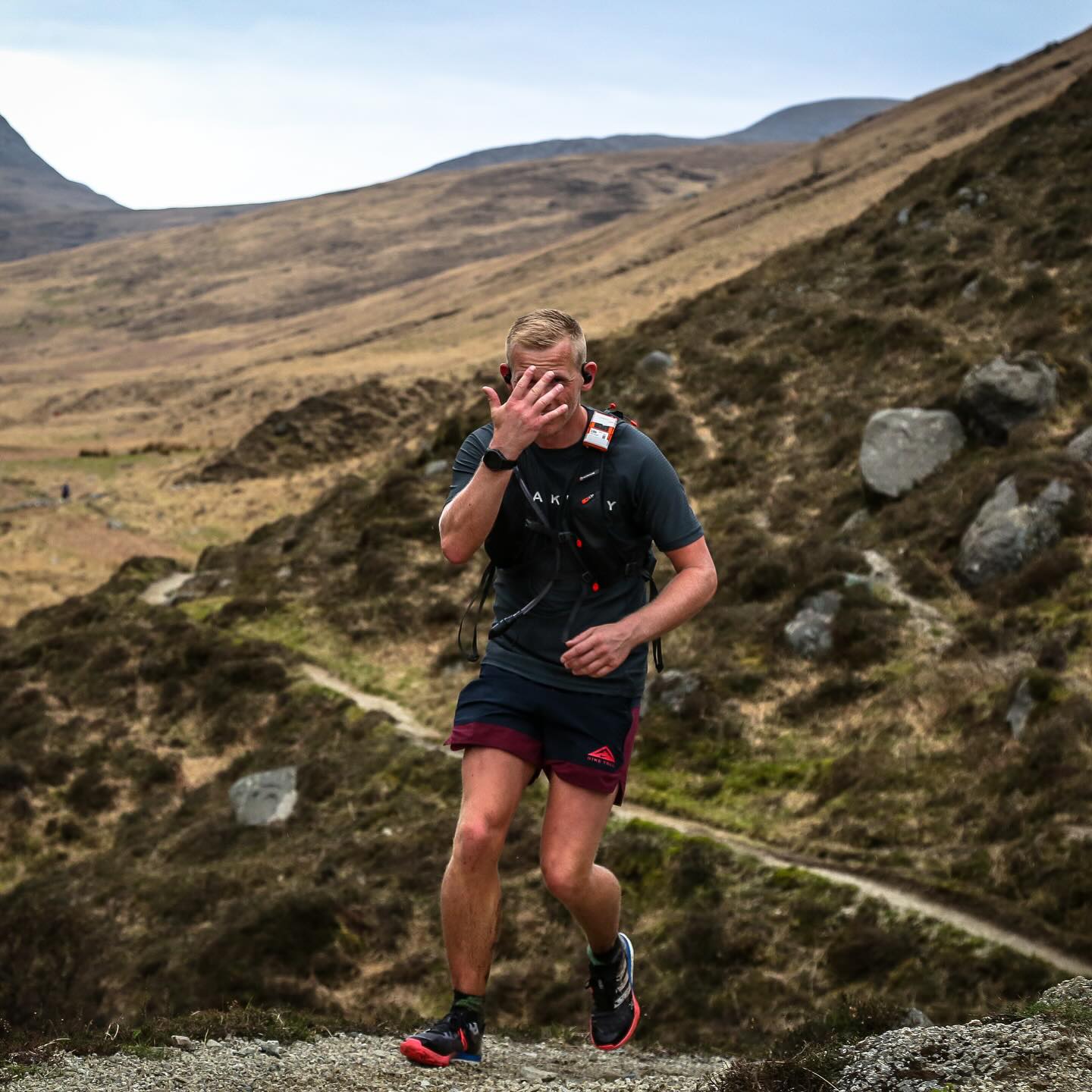 A runner climbing a rough hillside trail with a hand raised to his face.