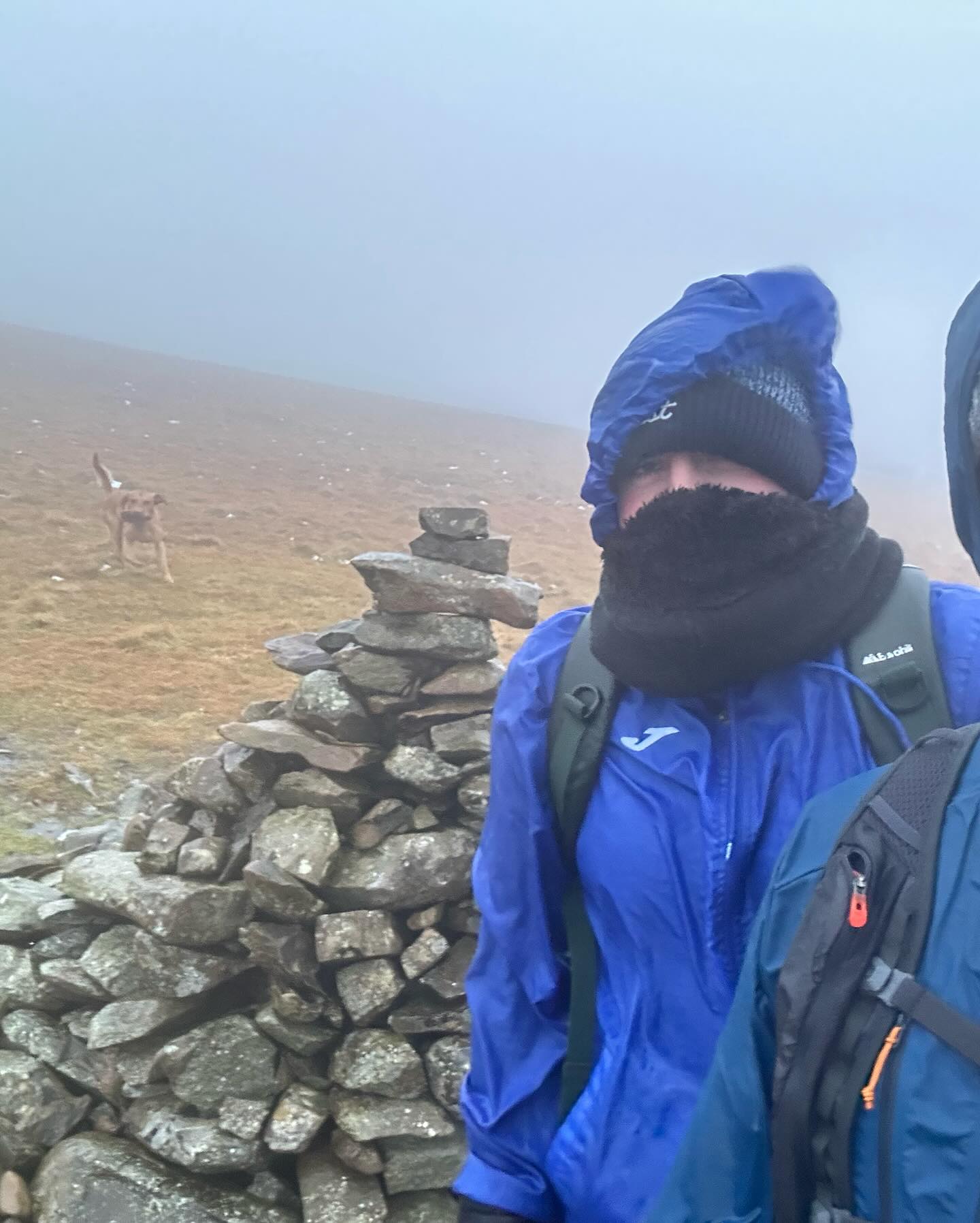 A walker in a blue jacket beside a stone cairn in mist.