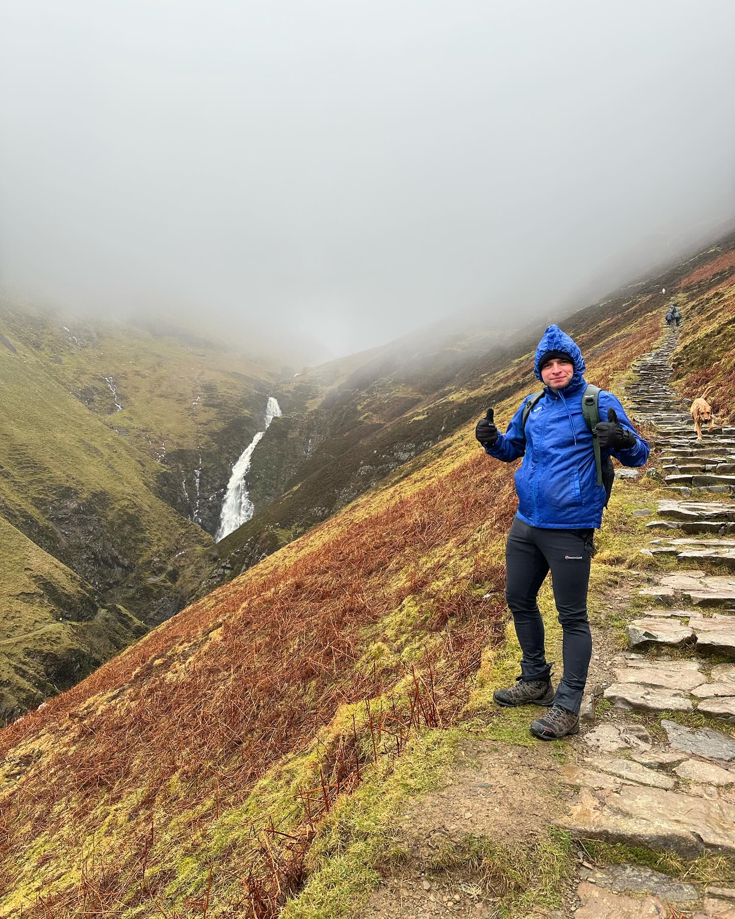 A walker on stone steps with a waterfall across the misty valley.