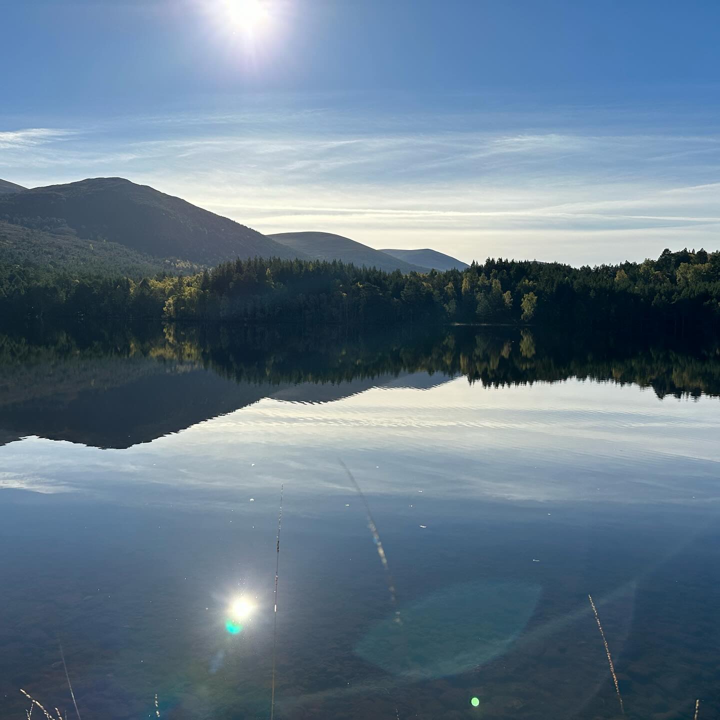Hills and trees reflected in the loch beneath bright sunshine.